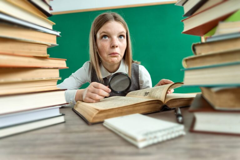 young female student between two stacks of books holding a magnifying glass above a book